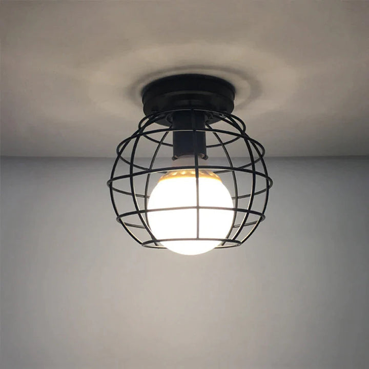 An industrial-style ceiling light with a globe cage design, featuring a exposed bulb, in a black color, placed above a white wall.