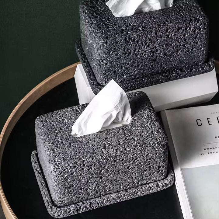 Black lava rock-style tissue box holder with textured stone finish, placed on a table beside a magazine.