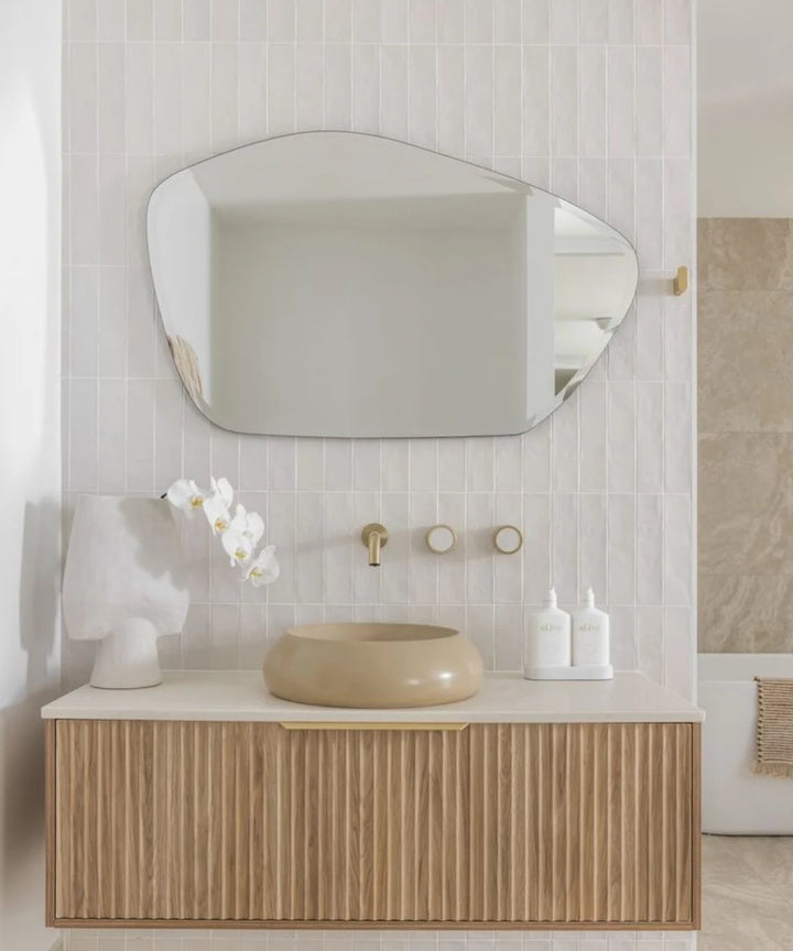 Asymmetrical modern wall mirror mounted above a beige vanity in a minimalist bathroom