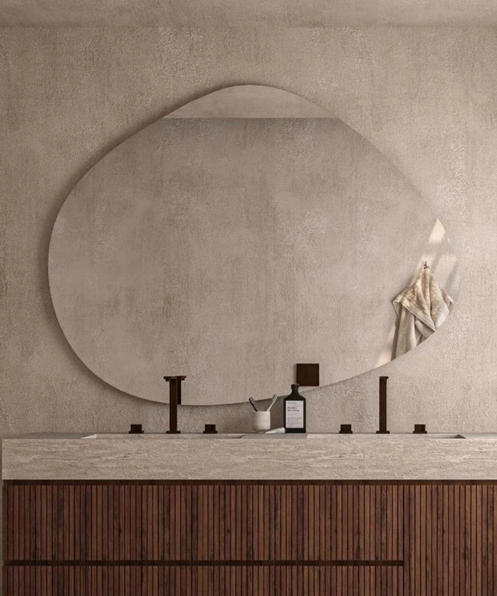 Organic modern wall mirror with a pebble-like shape above a dark wood vanity in a textured neutral-toned bathroom