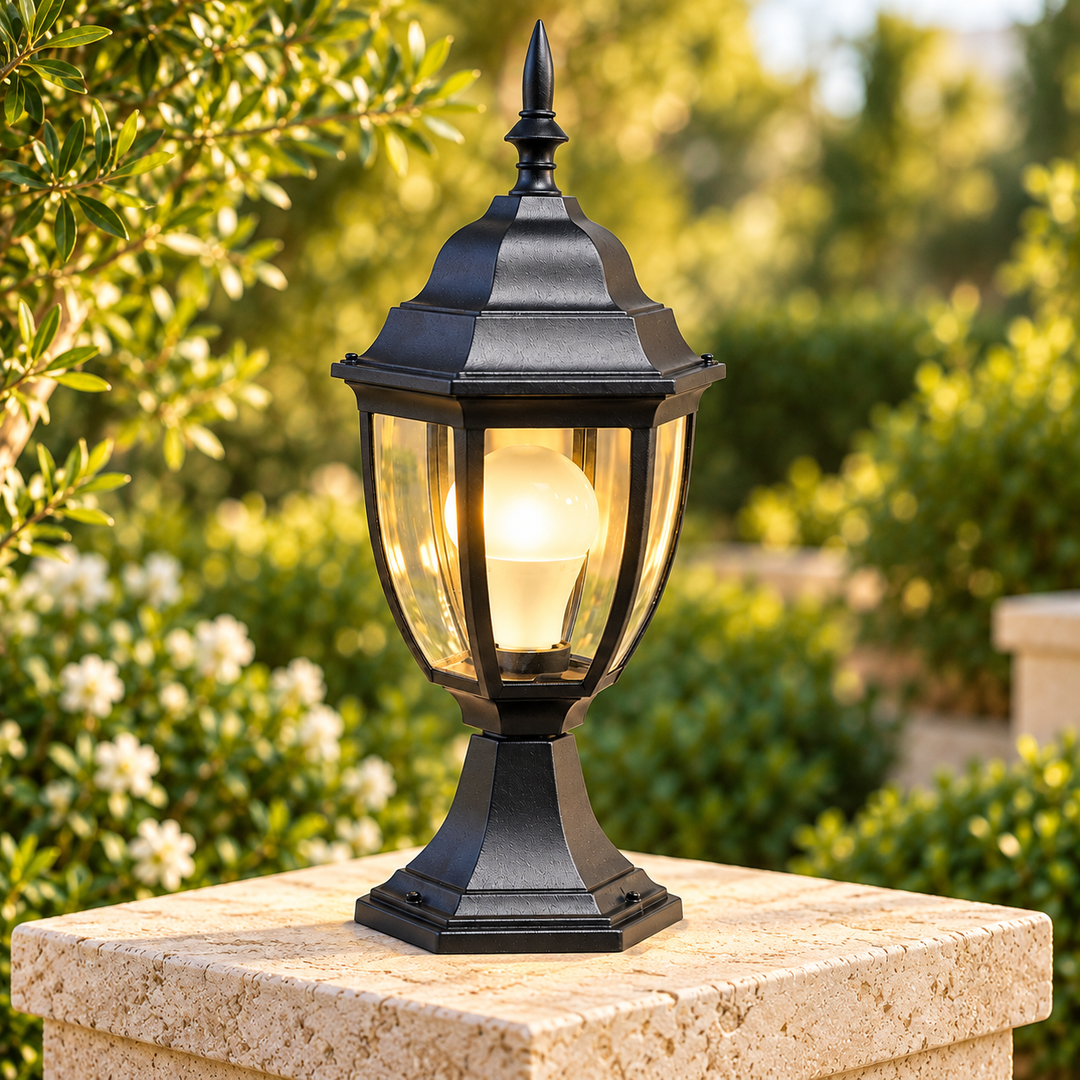 Traditional black outdoor lantern with clear glass panels and a warm glowing bulb displayed on a stone pedestal in a garden setting