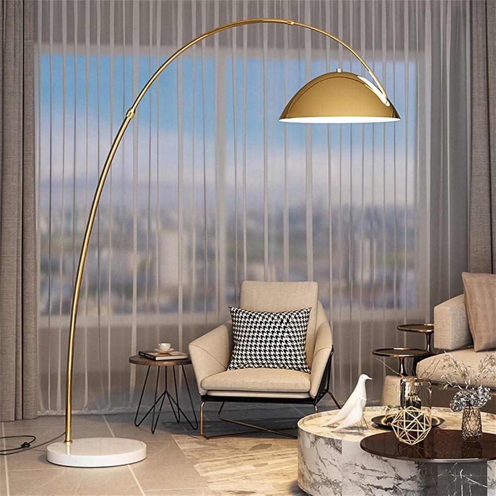Brass-tone arc floor lamp with a dome shade and round white marble base beside a lounge chair in a living room.