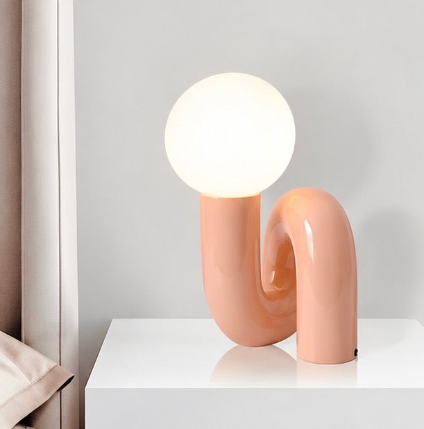 Sculptural peach table lamp with a curved loop-shaped base and glowing white globe shade on a white table