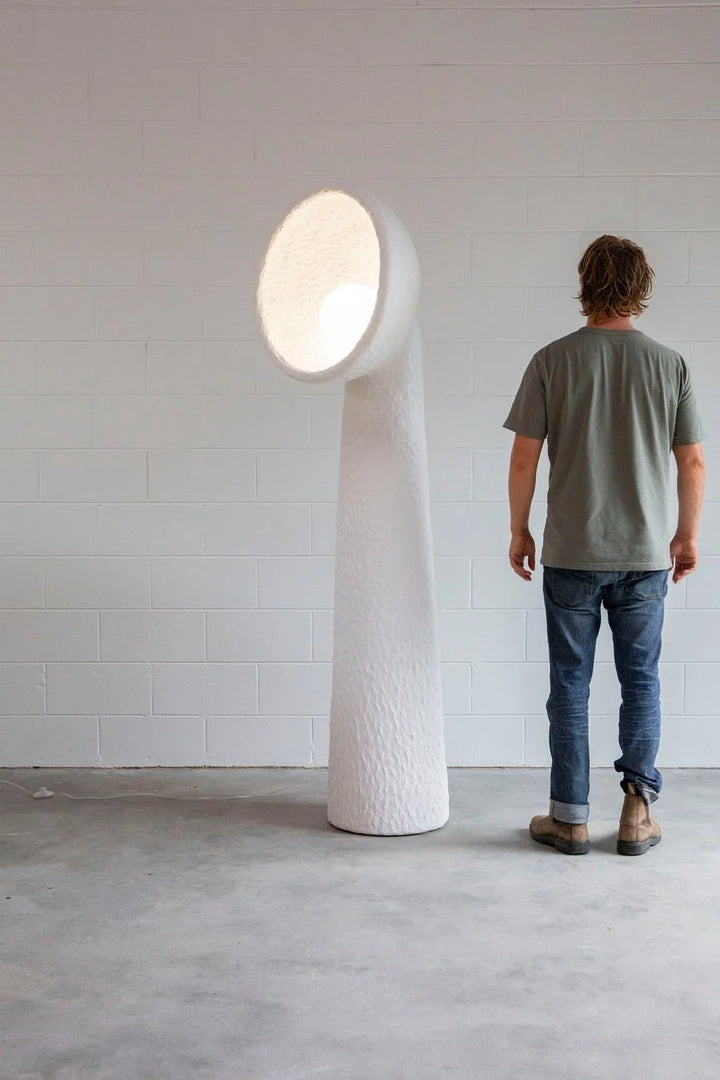 Tall white sculptural floor lamp with a curved textured base and glowing oval shade beside a person for scale