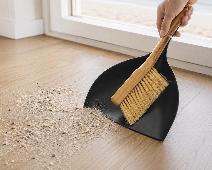 Black dustpan with natural wooden hand brush cleaning sand from wooden floor.