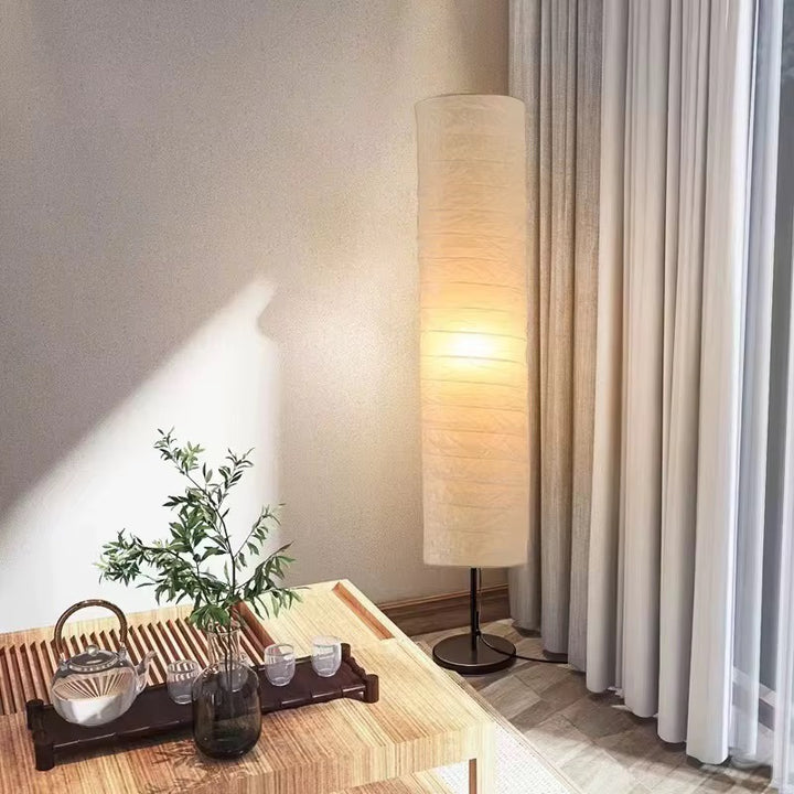 Tall beige cylindrical fabric floor lamp with a warm glow beside curtains and a dining table