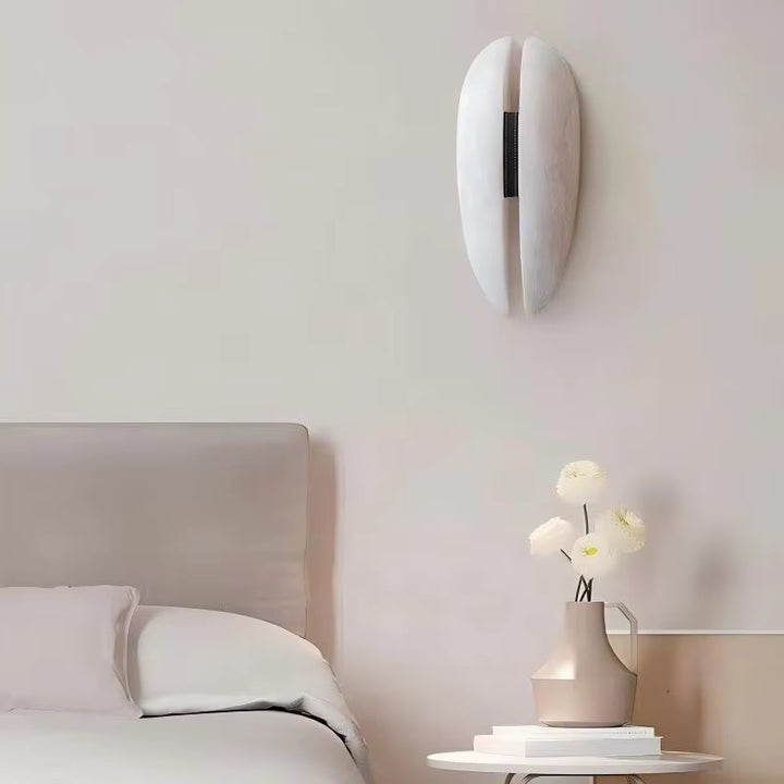 Sculptural white oval wall light with a narrow dark central detail above a bedroom nightstand