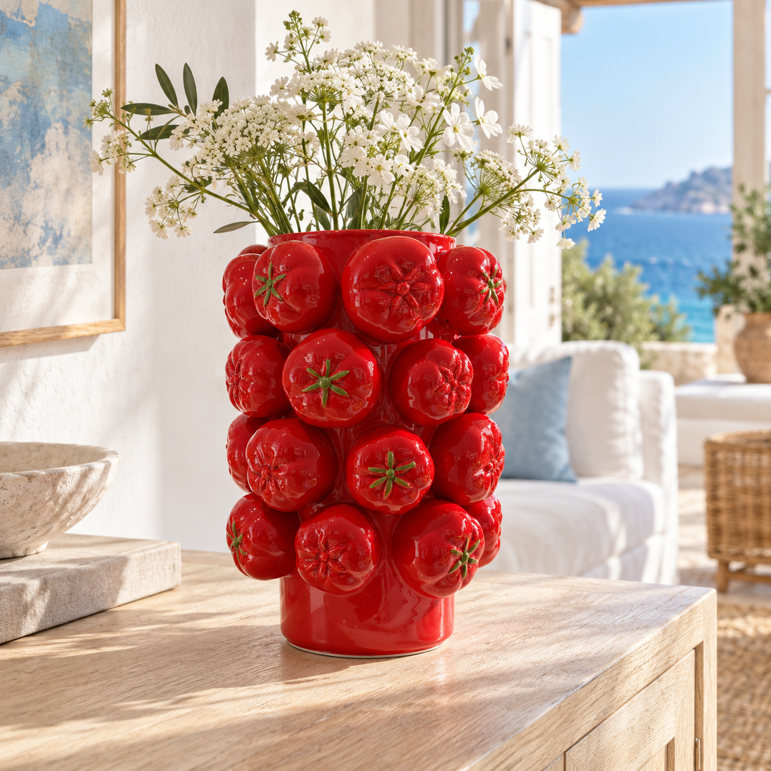 Bright red vase with a three-dimensional tomato design holding white flowers and greenery on a light wood table.