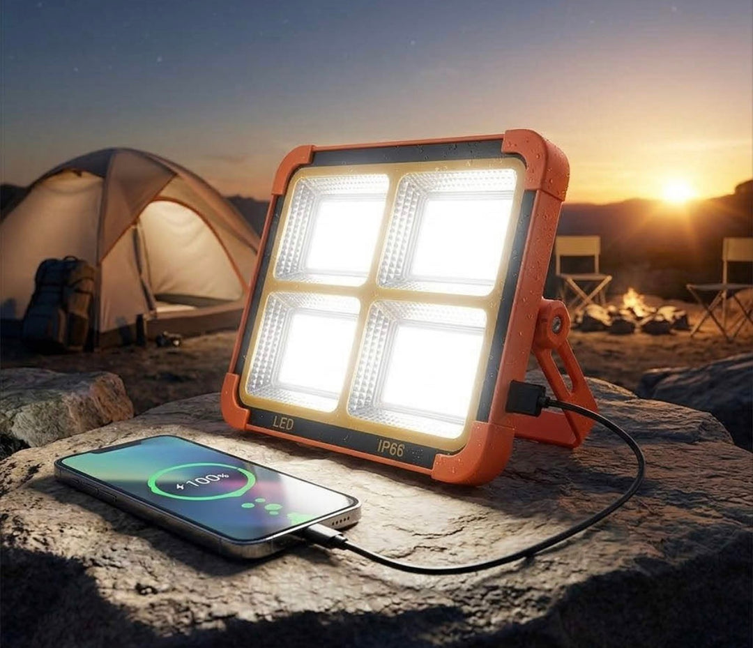 Orange portable LED floodlight with four illuminated panels beside a smartphone at a campsite