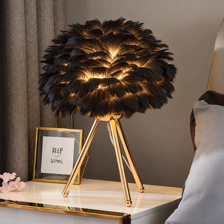 Black feather-style table lamp with a glowing shade and gold-tone tripod base on a bedside table