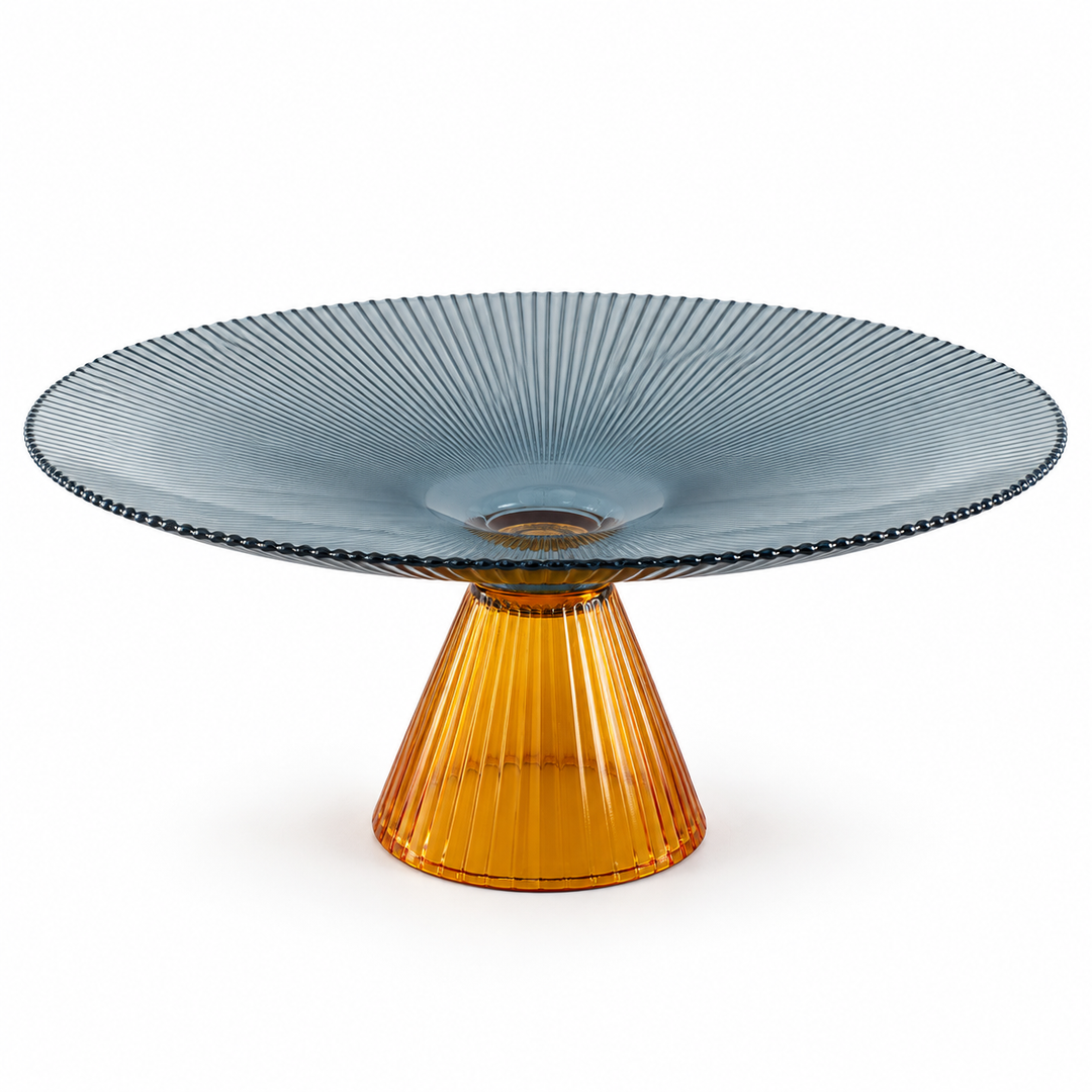 Wide glass fruit bowl with a dark patterned rim and fluted amber pedestal base, holding lemons, pears, and grapes on a dining table.