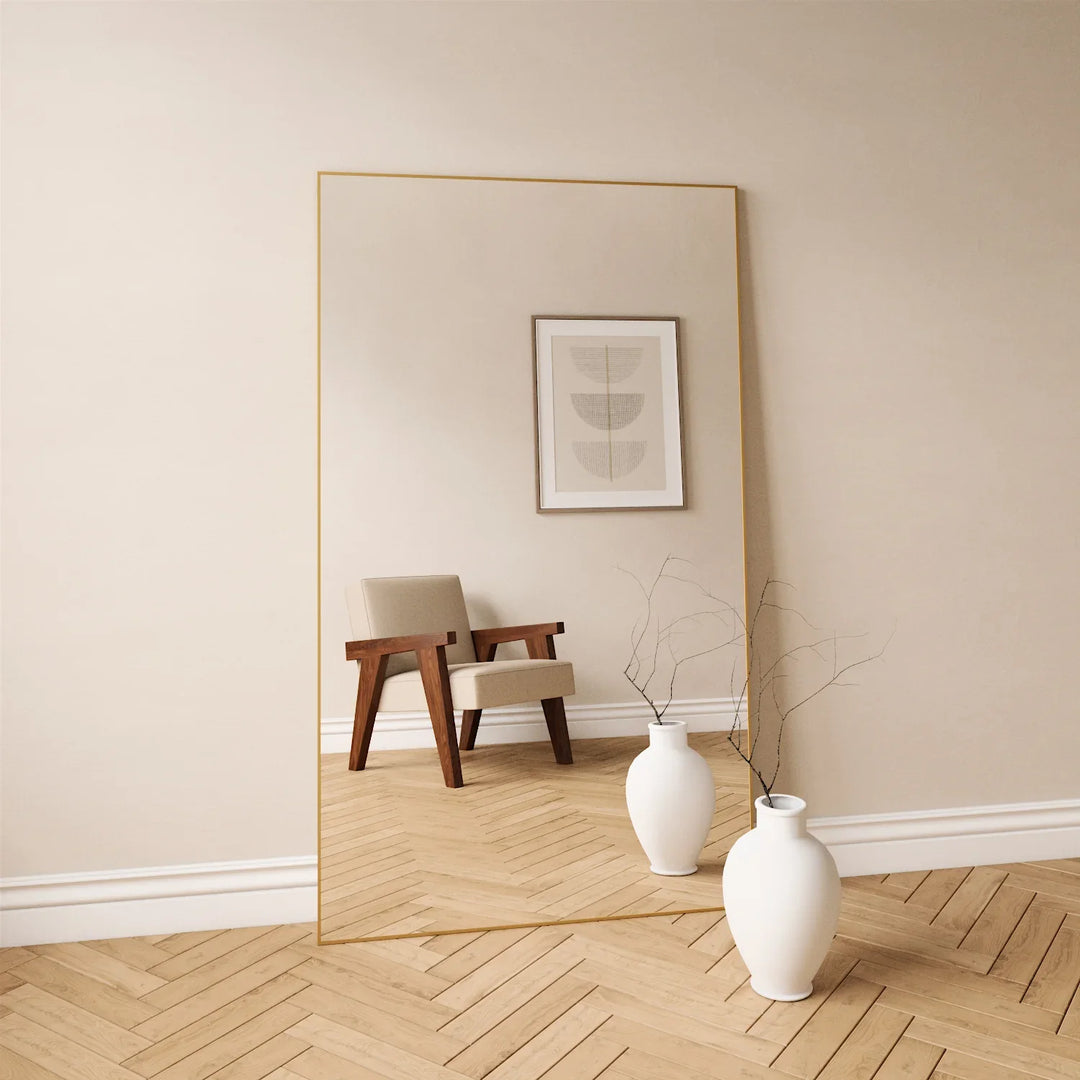 Full-length mirror with gold metal frame leaning on beige wall beside white vases