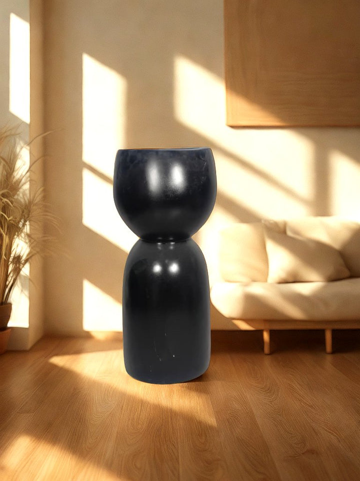 Glossy black fiberglass planter with a rounded bowl and tall sculptural pedestal-style base styled in a warm living room.