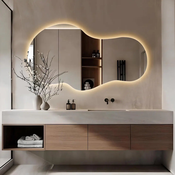 An asymmetrical large modern hanging mirror with a frameless design, featuring built-in lights, above a bathroom sink and vanity.