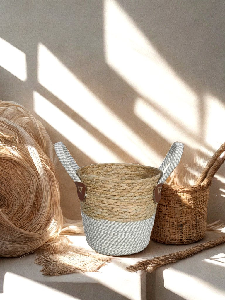 Natural woven basket planter with grey and white rope base and two loop handles, displayed with weaving materials in warm sunlight.
