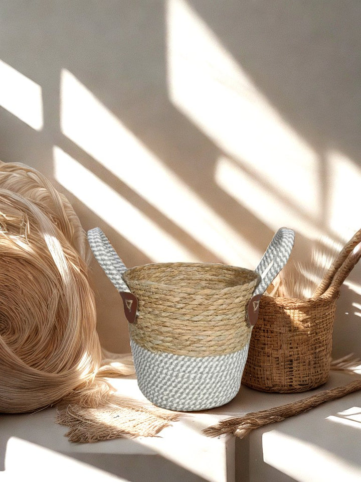 Natural woven basket planter with grey and white rope base and two loop handles, displayed with weaving materials in warm sunlight.