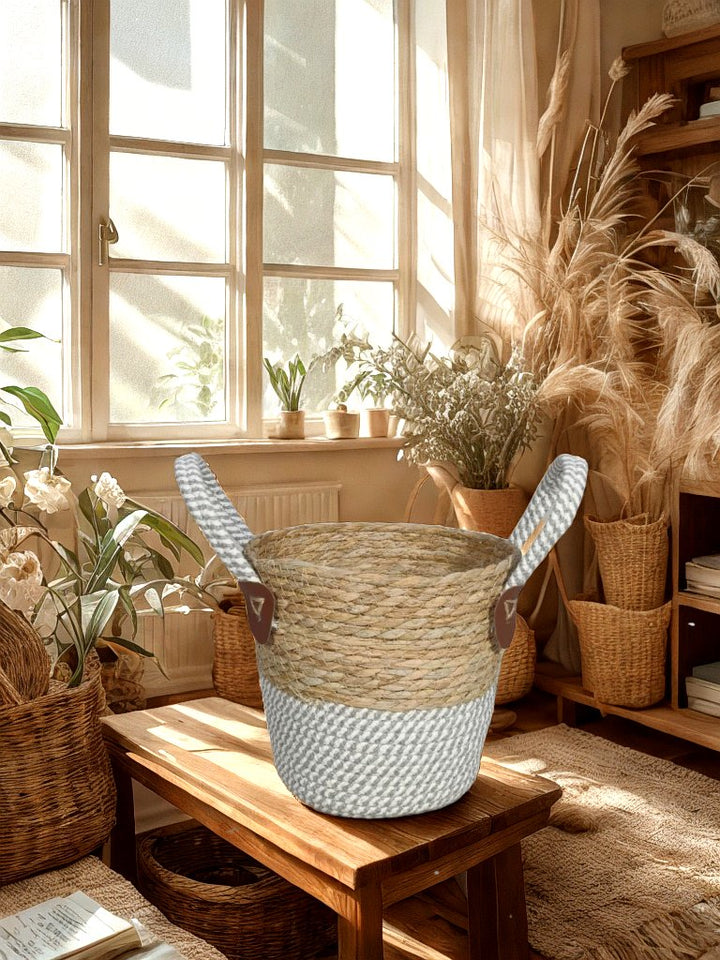 Natural and grey woven basket planter with two rope handles on a wooden stool in a sunlit room with decorative plants.