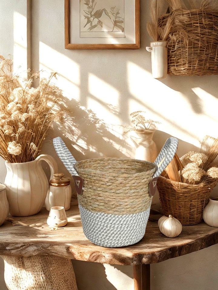 Two-tone woven basket planter with grey and white rope base and handles on a wooden table beside neutral vases and dried flowers.