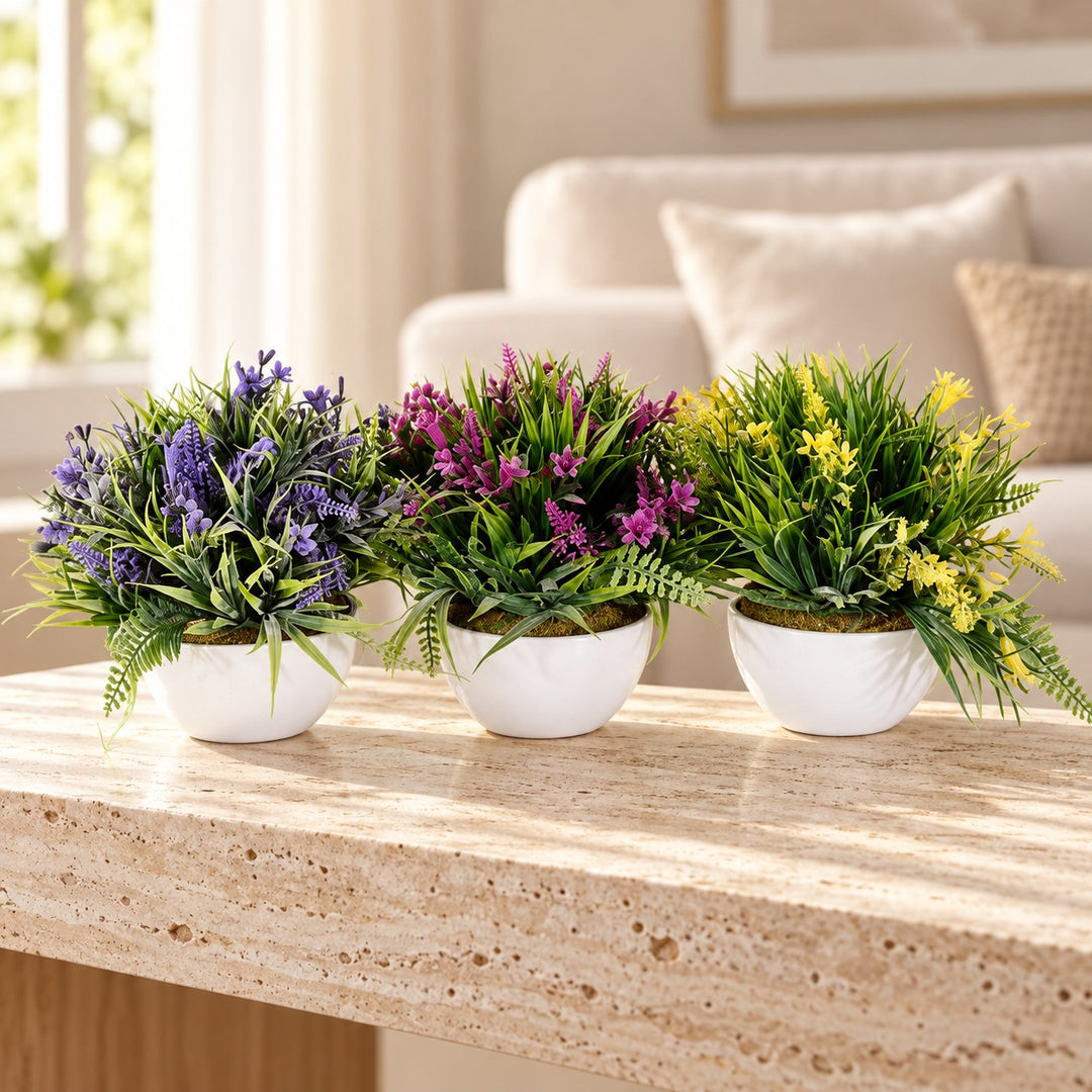 Three artificial flowering plants in purple, magenta and yellow with green foliage in rounded white pots styled on a travertine table in an elegant neutral living room