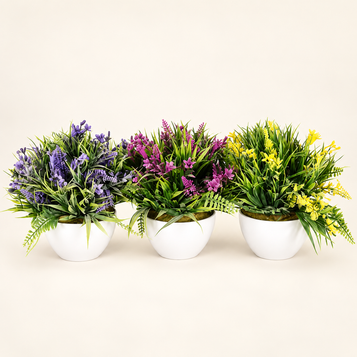 Three artificial flowering plants in purple, magenta and yellow with green foliage in rounded white pots on an ivory studio background
