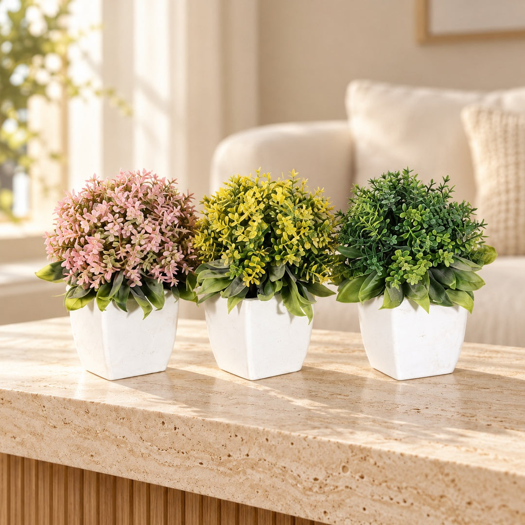 Set of three rounded artificial plants with pink, yellow and green foliage in white faceted pots styled on a travertine table in an elegant neutral living room