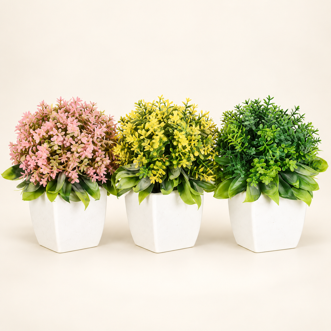 Set of three rounded artificial plants with pink, yellow and green foliage in white faceted pots on an ivory studio background