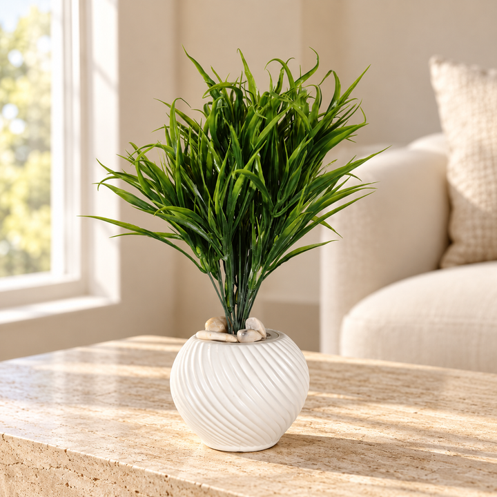 Artificial green grass with curling blades in a round white spiral-ribbed pot topped with pale stones styled on a travertine table in an elegant neutral living room