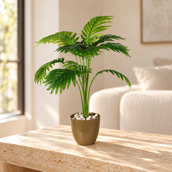 Artificial palm with arching green fronds in an olive-gold pot topped with pale decorative stones styled on a travertine table in an elegant neutral living room