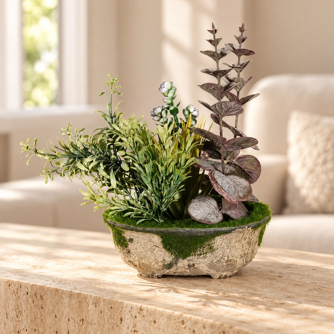 Artificial arrangement with muted eucalyptus-style leaves, green meadow foliage and moss in a rustic oval planter styled on a travertine table in an elegant neutral living room