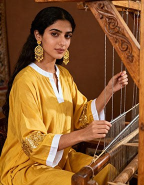 Wadi Home framed canvas print of a woman weaving traditional fabric. She wears a yellow tunic with gold embroidery and white cuffs, large gold earrings.