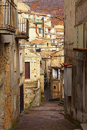 Wadi Home European village street scene wall art, sun-drenched alley with rustic buildings and balconies.