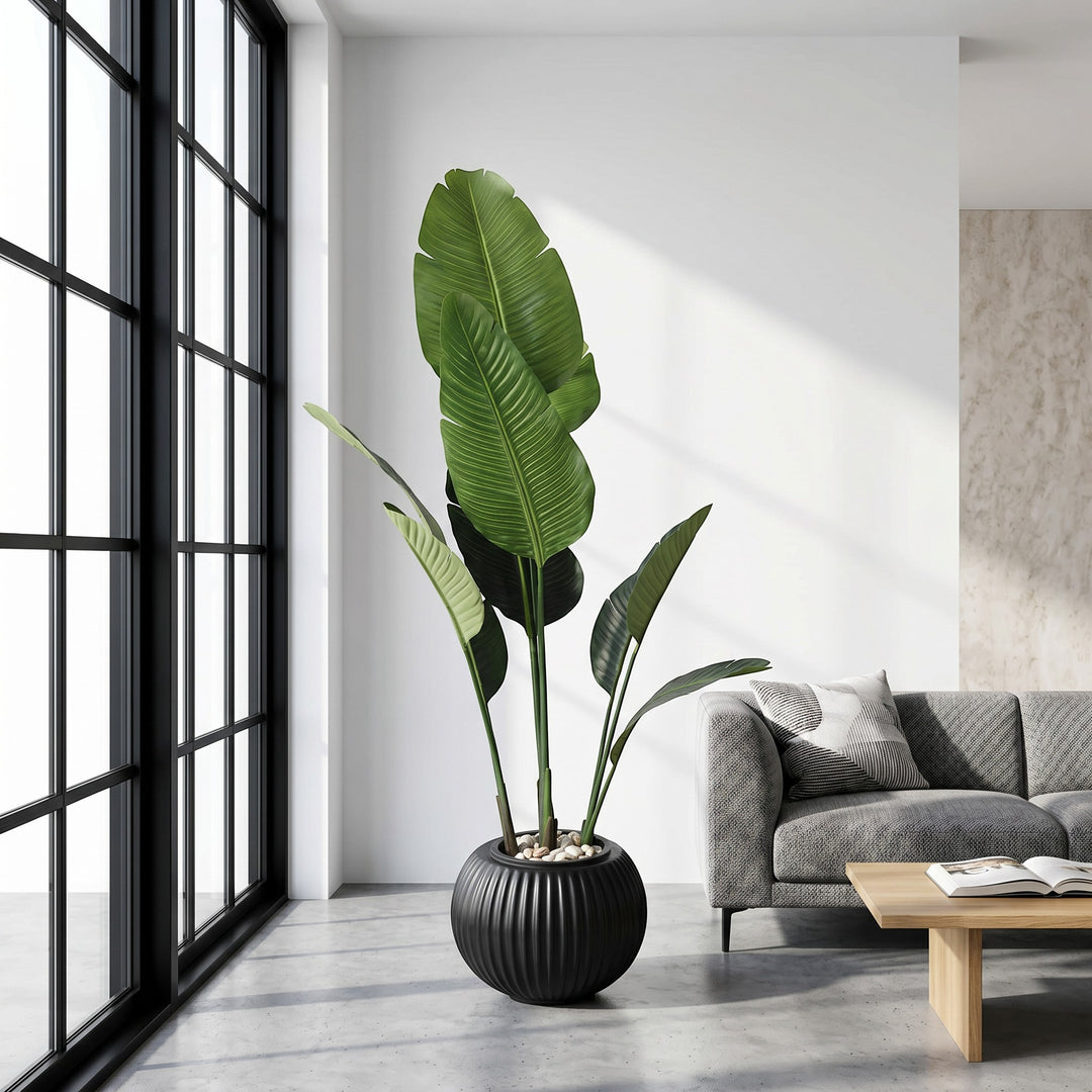 Wadi Home large artificial Bird of Paradise plant in a black ribbed planter with white stones, next to a grey sofa.