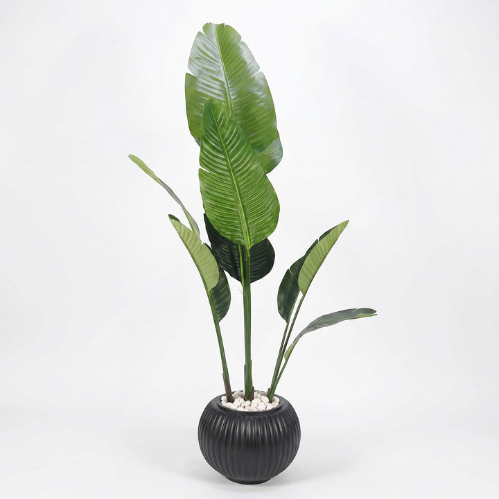 Wadi Home artificial Bird of Paradise plant with large green leaves in a fluted black pot with white stones.