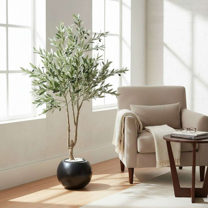 Wadi Home artificial olive tree with green leaves and black olives in a round black planter, placed in a living room.