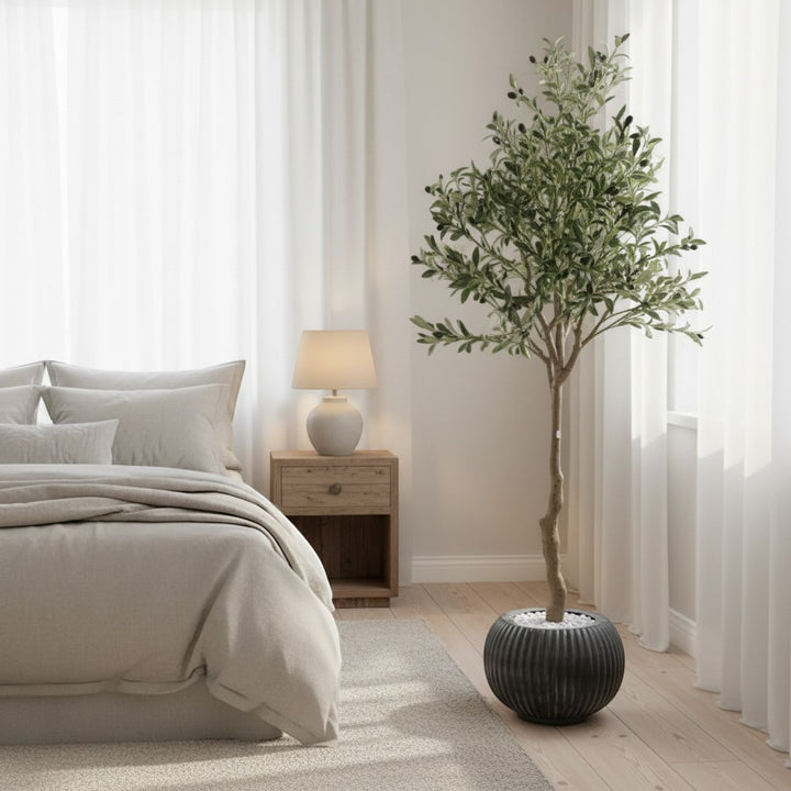 Wadi Home artificial olive tree with green leaves in a ribbed black pot, placed in a modern neutral bedroom.