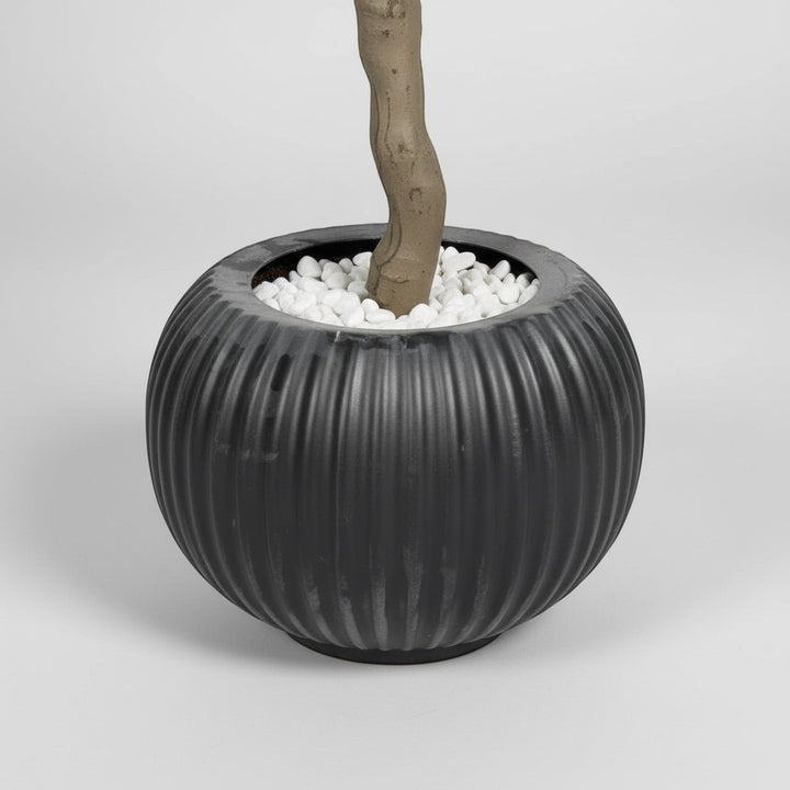 Wadi Home artificial olive tree in a round, ribbed black pot filled with decorative white pebbles.