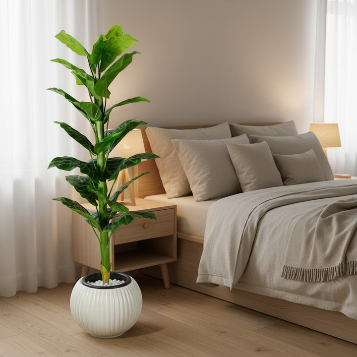 Wadi Home artificial floor plant with lush green leaves in a white ribbed decorative planter in a cozy bedroom.