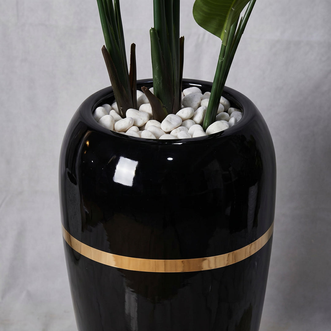 Wadi Home Large Bird of Paradise artificial plant in a tall black pot with a gold band and white stones.