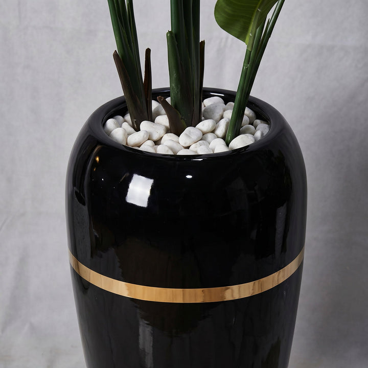 Wadi Home Large Bird of Paradise artificial plant in a tall black pot with a gold band and white stones.
