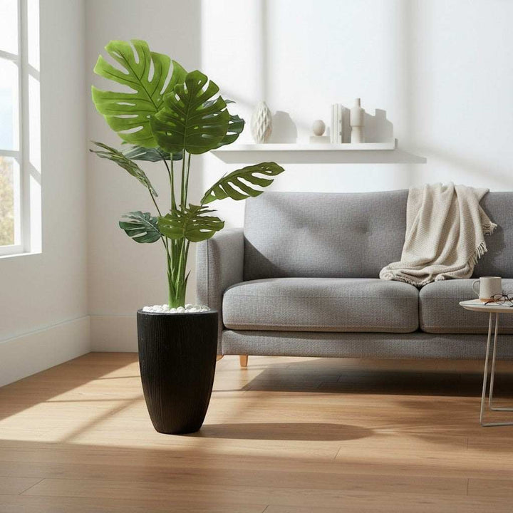 Wadi Home Large artificial Monstera plant with split green leaves in a textured black pot with white stones.