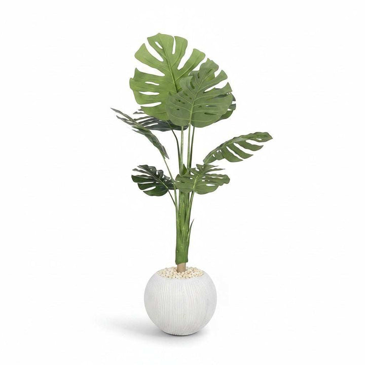 Wadi Home large artificial Monstera plant with deep green split leaves in a textured white spherical pot with decorative stones.