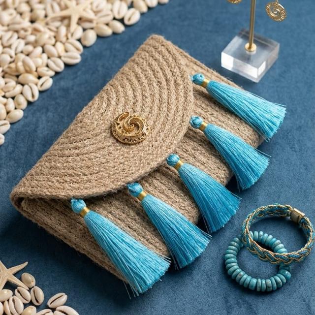Wadi Home woven jute clutch handbag featuring turquoise tassels and a gold-tone metal accent on a blue background.