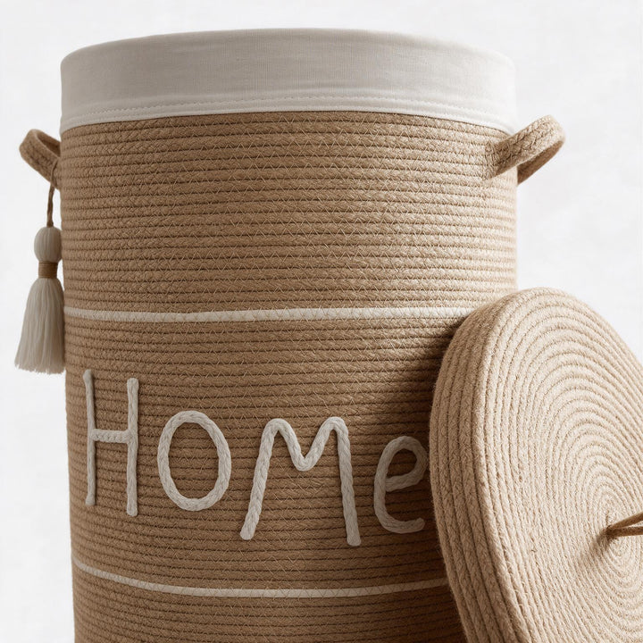 Wadi Home woven rope laundry hamper in beige with a white lid, featuring a decorative Home script and side tassels.