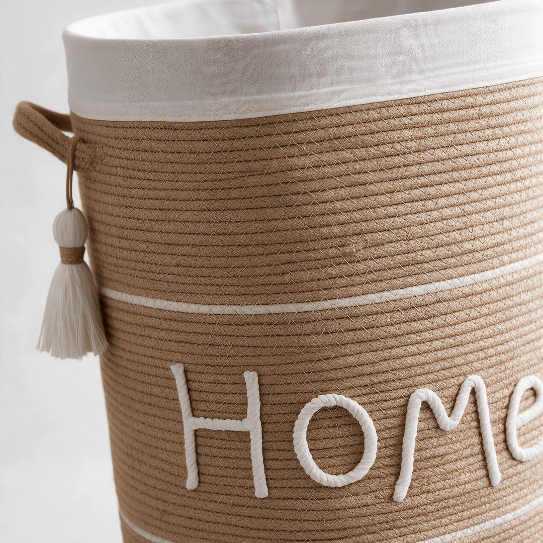 Wadi Home woven rope laundry hamper in beige with white Home lettering, tassel detail, and white fabric lining.