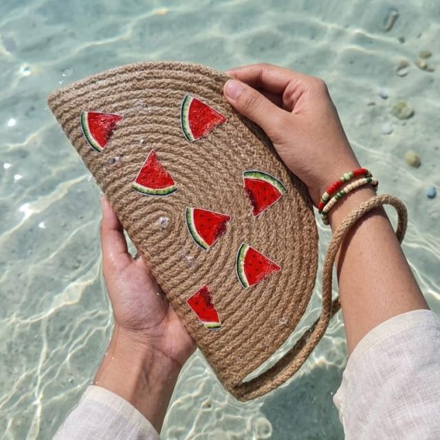 Wadi Home beige woven half moon clutch with playful watermelon slice prints, held over clear beach water.