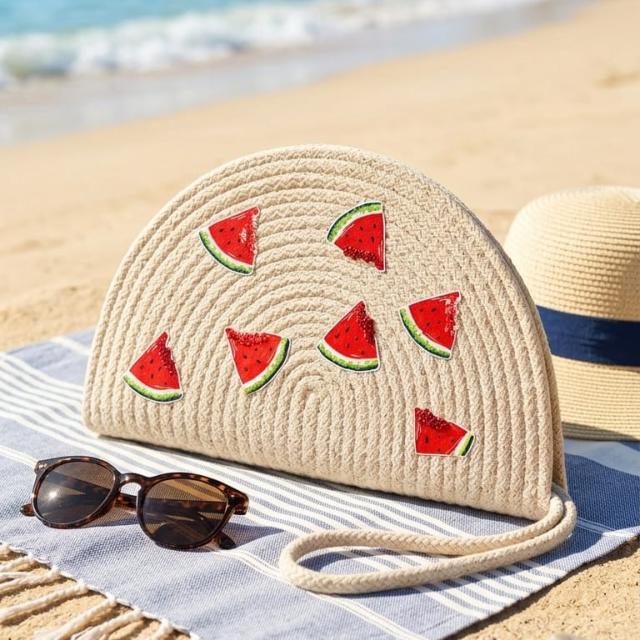 Wadi Home beige woven half moon clutch with playful watermelon slice prints, displayed on a beach towel.