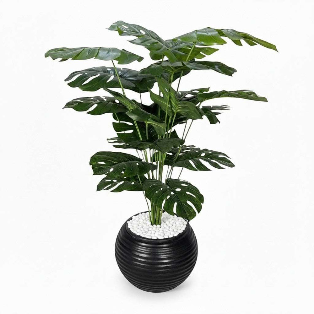 Wadi Home large artificial Monstera plant with deep green fenestrated leaves in a black ribbed planter with white stones.